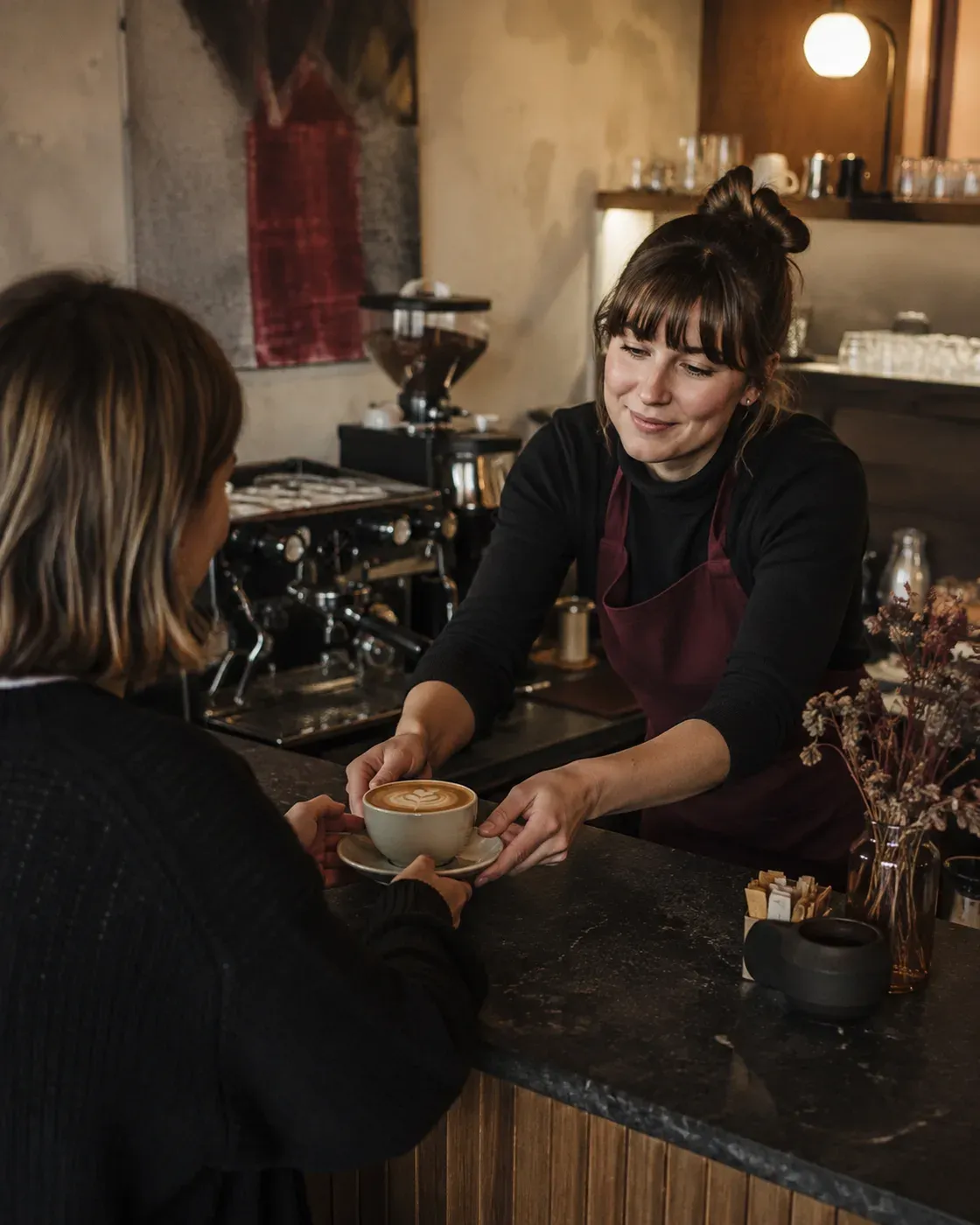 Illustrative scene of a barista handing coffee to a guest at the espresso bar, with the grinder and coffee beans in view.