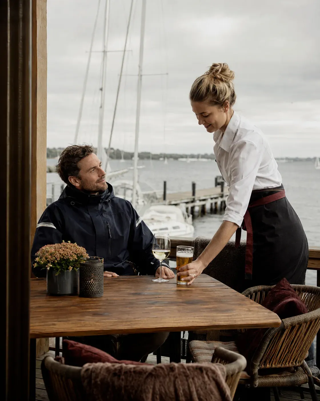 Illustrative scene of a team member bringing drinks to sailors on a marina terrace, with the jetty and boat masts behind them.